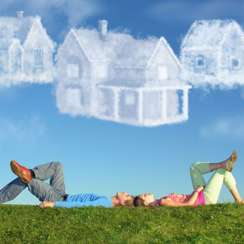 A whimsical image of a man and woman lying back-to-back on a grassy hill, gazing upwards at clouds shaped like various styles of houses. The sky is clear and blue, creating a stark contrast with the white, fluffy cloud houses above them.