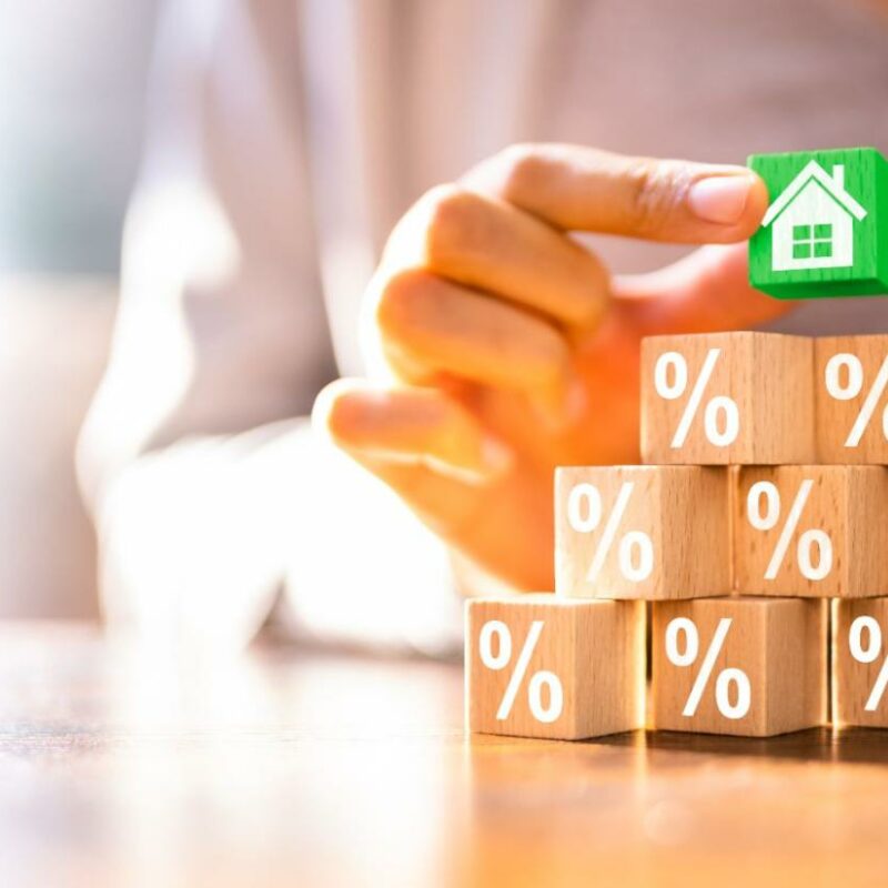 A person's hand placing a small, green house figurine on top of stacked wooden blocks, each marked with a percentage symbol, symbolizing mortgage rates or financing options.