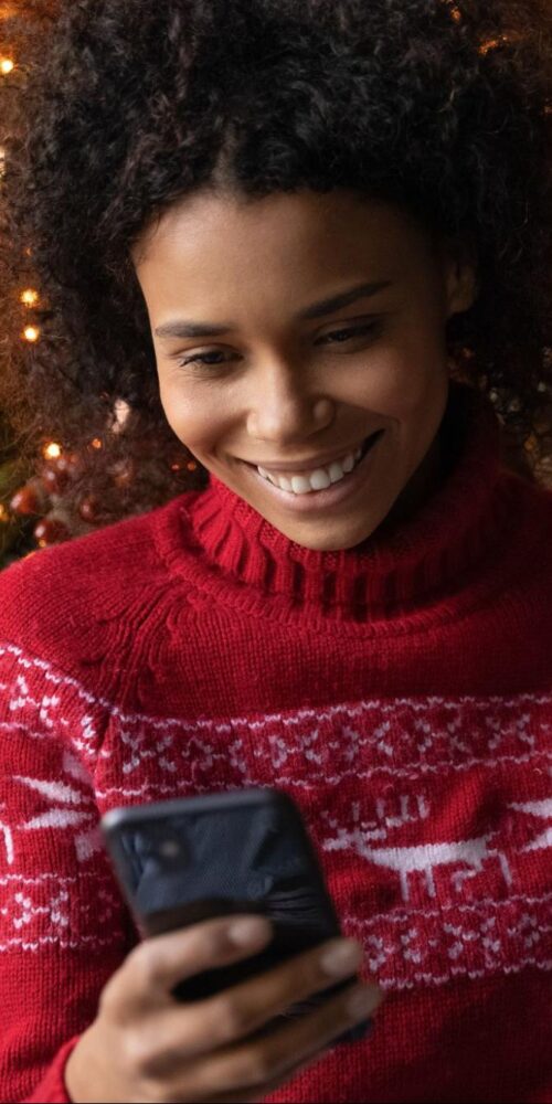 Woman smiling while holiday shopping online, holding a credit card in front of a decorated Christmas tree.