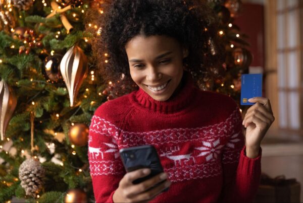 Woman smiling while holiday shopping online, holding a credit card in front of a decorated Christmas tree.