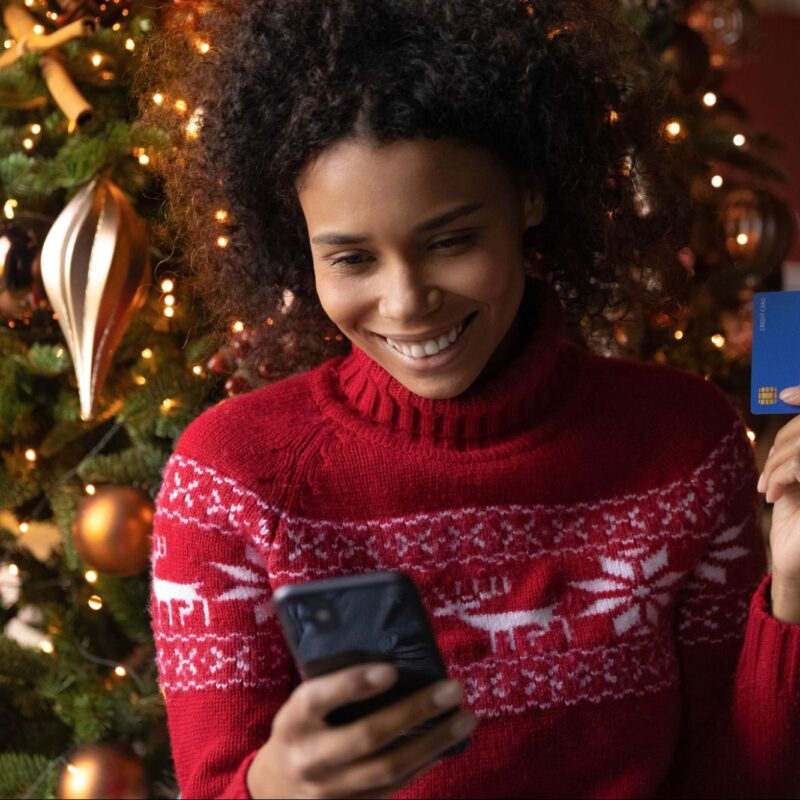 Woman smiling while holiday shopping online, holding a credit card in front of a decorated Christmas tree.